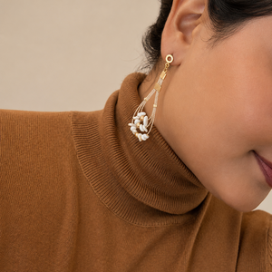A close-up of a woman wearing a gold and white beaded dangle earring, paired with a brown ribbed turtleneck sweater against a neutral beige background.