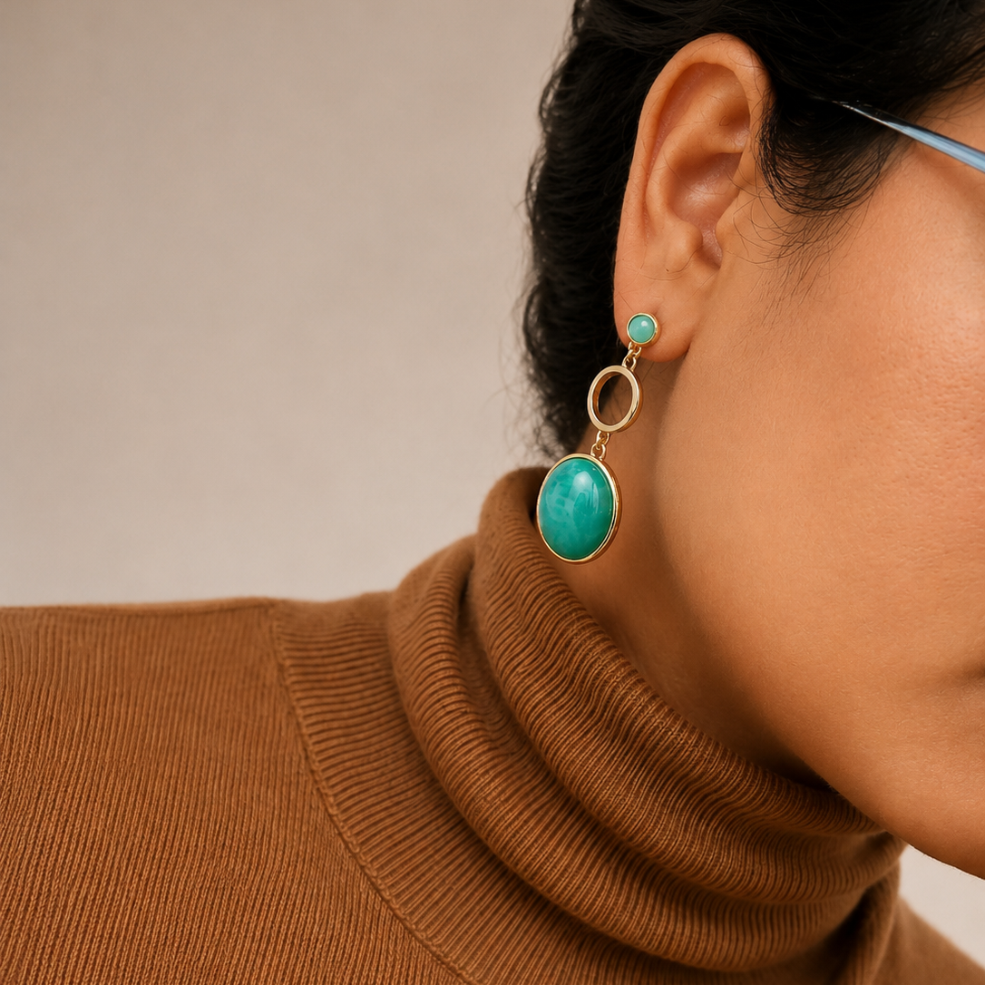 A close-up of a model wearing a gold drop earring with a large oval turquoise stone and a matching small stud earring, paired with a brown ribbed turtleneck sweater.